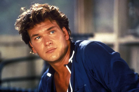 Patrick Swayze as Johnny Castle, a light-skinned man with short, styled brown hair wearing a casual blue button-down hair is looking off camera.